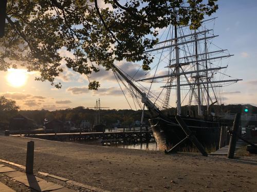 A historic ship docked by a waterfront at sunset, with trees and a calm sky in the background.