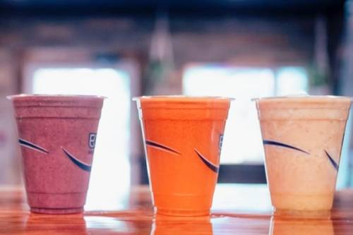 Three colorful smoothie cups in a row: purple, orange, and beige, on a wooden table with a blurred background.