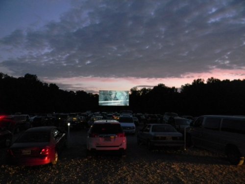 A drive-in movie theater at dusk, with cars parked and a large screen displaying a film against a cloudy sky.