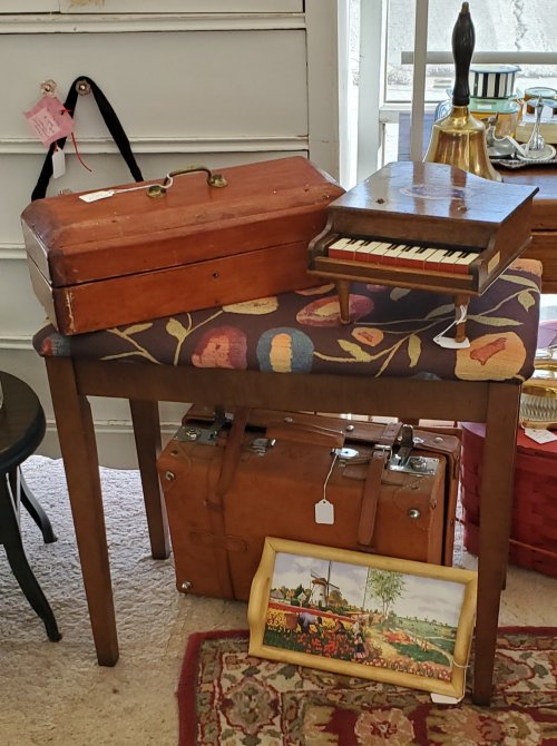 A wooden table with a floral-patterned top, featuring a small piano, a toolbox, and vintage suitcases underneath.