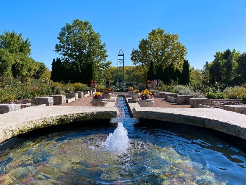 A serene garden scene featuring a fountain, flower beds, and a clock tower under a clear blue sky.