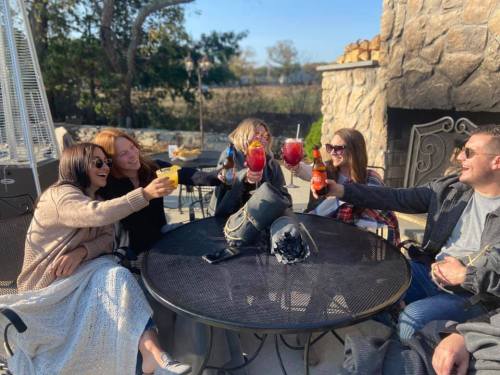 A group of five friends toasting with colorful drinks around a table outdoors, enjoying a sunny day.