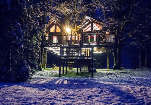 A cozy treehouse illuminated at night, surrounded by snow-covered ground and trees.