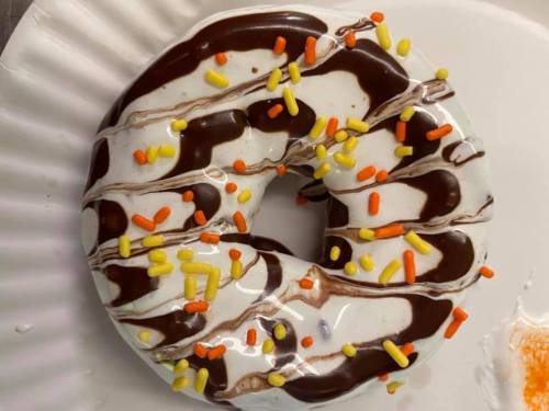 A frosted donut with chocolate drizzle and colorful sprinkles on a paper plate.