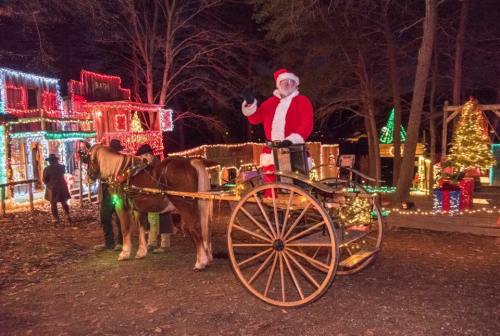 A festive scene with Santa in a sleigh, surrounded by colorful holiday lights and decorations in a wooded area.