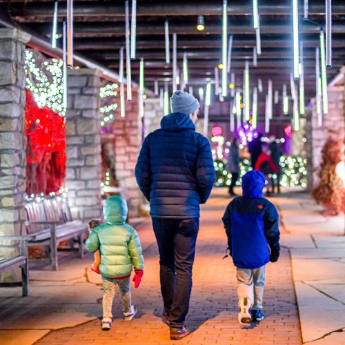 A person walks with two children through a festive, illuminated pathway lined with colorful lights.
