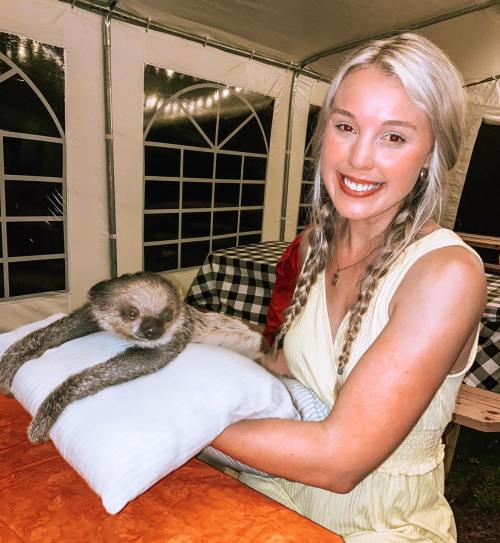 A smiling woman holds a sloth on a pillow while sitting at a table in a tent.