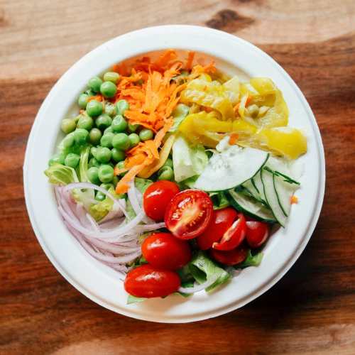 A colorful salad bowl with greens, cherry tomatoes, cucumbers, carrots, peas, and yellow peppers on a wooden surface.