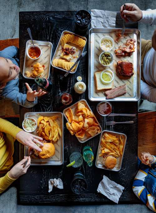 A top-down view of a table filled with various BBQ dishes, sides, and drinks, with hands reaching for food.