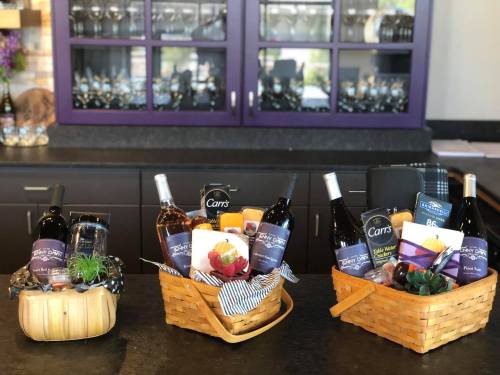 Three gift baskets filled with wine, snacks, and decorative items, displayed on a countertop with glassware in the background.