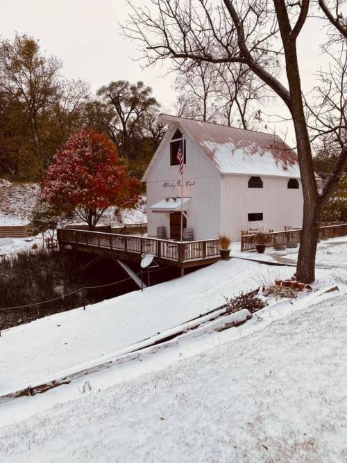 A quaint white barn with a porch, surrounded by snow and autumn trees, featuring an American flag.