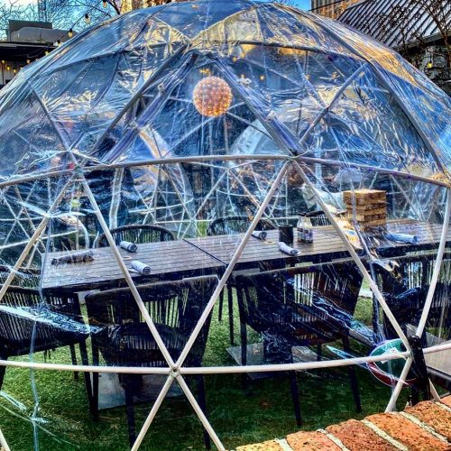 A transparent geodesic dome with a dining table and chairs inside, set on a grassy area.