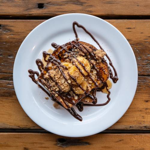 A plate of dessert featuring banana slices, chocolate drizzle, and crumbled toppings on a wooden table.