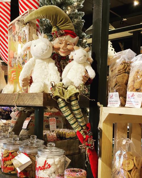 A festive elf with a green hat holds two white stuffed bunnies, surrounded by holiday treats and decorations.