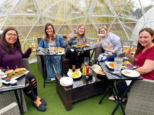 A group of five women enjoying a meal inside a clear dome, smiling and raising drinks with various dishes on the table.