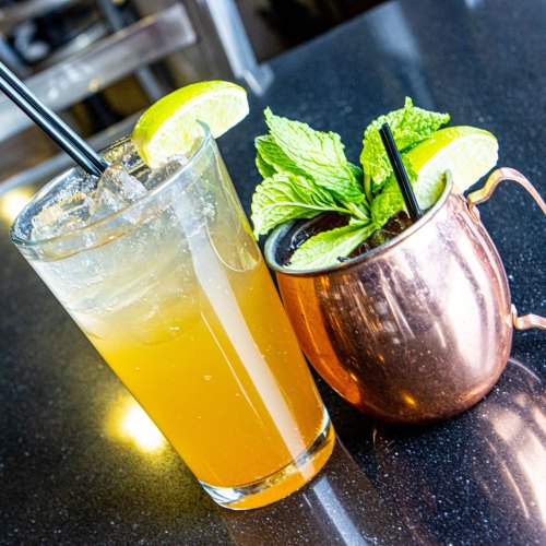 Two cocktails on a black table: a tall glass with a citrus drink and a copper mug filled with a dark beverage and mint.