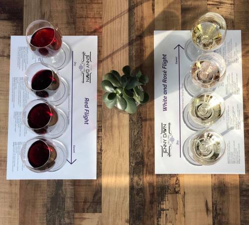 Two flight trays of wine glasses on a wooden table, featuring red wines on the left and white/rosé wines on the right.