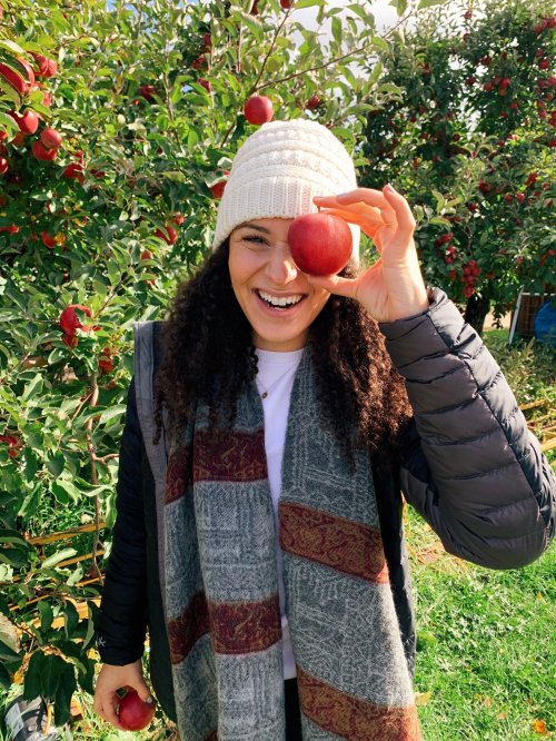 A smiling person wearing a beanie and scarf holds an apple in front of their face, surrounded by apple trees.