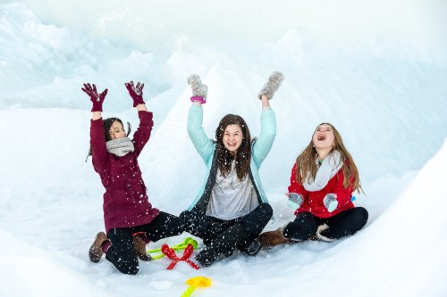 Three young women sitting in snow, joyfully throwing snow in the air, surrounded by icy formations.