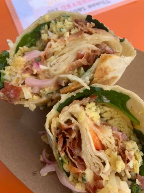 A close-up of a halved wrap filled with spinach, bacon, eggs, cheese, and tomatoes on a brown tray.