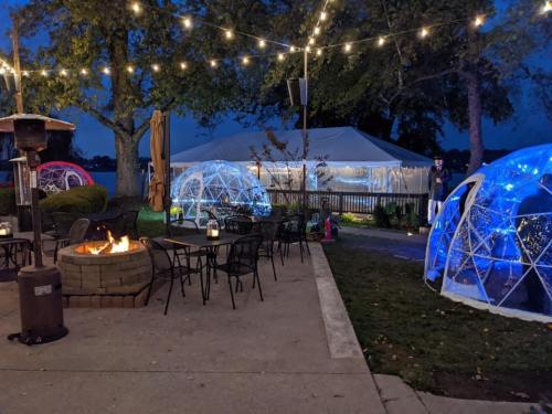 Outdoor seating area with glowing domes, a fire pit, and string lights at dusk. A tent is visible in the background.