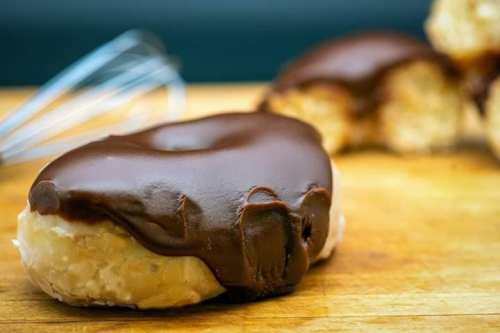 A close-up of a chocolate-glazed donut on a wooden surface, with a whisk in the background.