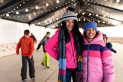 Two girls in winter coats and hats smile while ice skating indoors, with other children skating in the background.