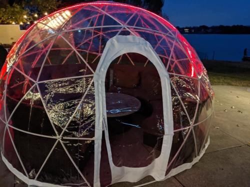 A clear geodesic dome with red lights, featuring a cozy seating area, set by a lake at night.