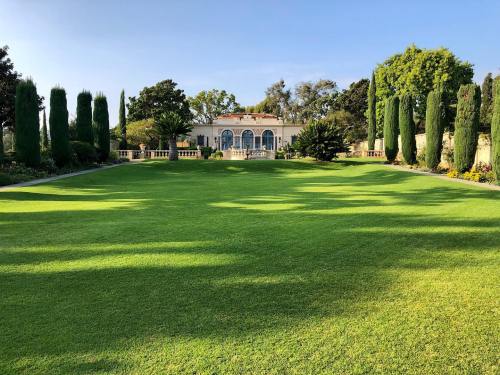 A lush green lawn leads to a grand building surrounded by tall trees and manicured hedges under a clear blue sky.