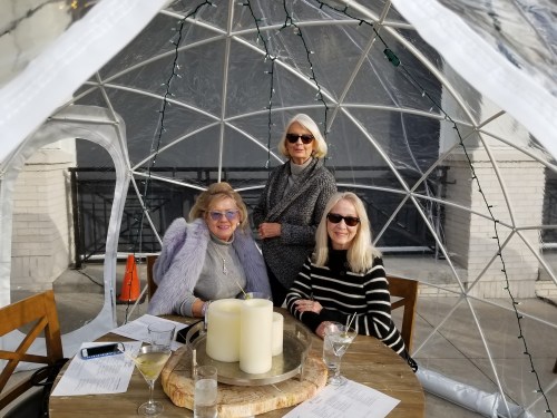 Three women sit at a table inside a clear igloo-like structure, enjoying drinks and smiling.