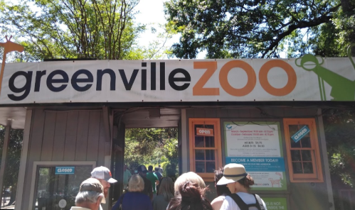 Entrance to Greenville Zoo with a large sign and visitors approaching the gate on a sunny day.
