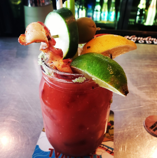 A vibrant Bloody Mary in a jar, garnished with bacon, cucumber, olives, and citrus slices on a metallic surface.