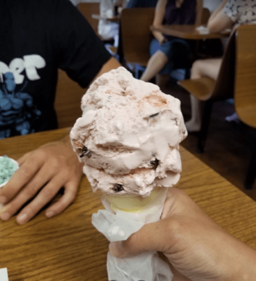 A hand holding a large scoop of pink ice cream on a cone, with people seated in the background.