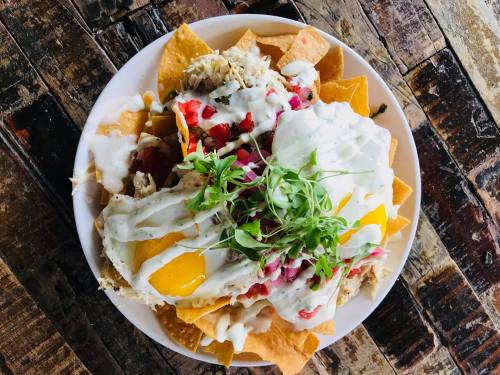 A plate of nachos topped with chicken, eggs, diced tomatoes, onions, and drizzled with creamy sauce, served on a wooden table.