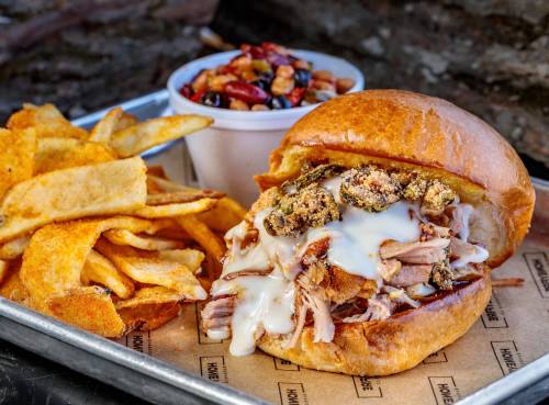 A pulled pork sandwich with creamy sauce, served with fries and a side of mixed nuts in a cup.