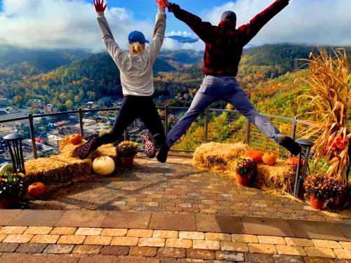 Two people jump joyfully in front of a scenic mountain view, surrounded by pumpkins and autumn decorations.