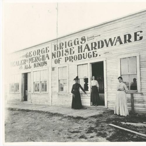 Historic black-and-white photo of a hardware store with three women, one standing outside and two inside.