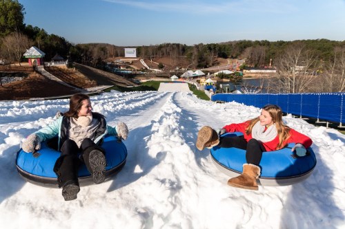 Two girls sit in snow tubes on a snowy slope, smiling at each other, with a winter amusement park in the background.