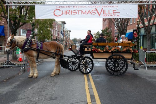 A horse-drawn carriage decorated for Christmas, with passengers, passes under a festive banner in a town street.