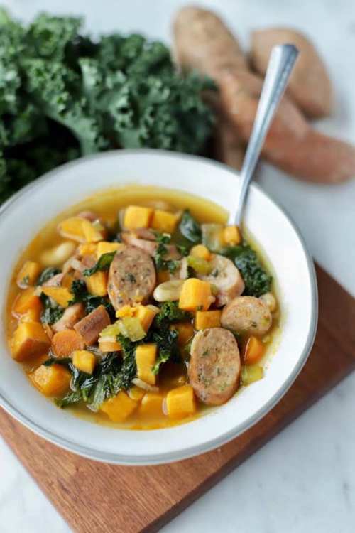 A bowl of hearty soup with sausage, sweet potatoes, kale, and vegetables, garnished with a spoon, on a wooden board.