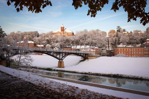A snowy landscape featuring a bridge over a river, with a historic building and trees in the background.