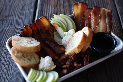 A platter featuring crispy bacon, sliced bread, green apple slices, and a small bowl of sauce on a wooden table.