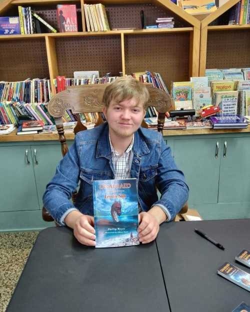 A young man in a denim jacket sits at a table holding a book titled "Unafraid: Jesus' Son" in a library setting.