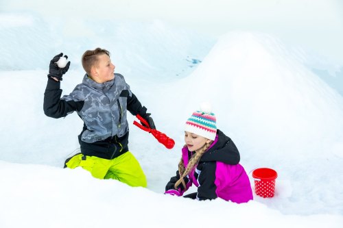 Two children playing in the snow: one is throwing a snowball while the other is sitting and building with snow.