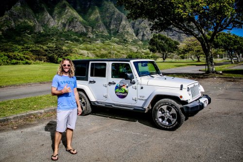 A man with long hair and sunglasses stands next to a white Jeep in a scenic park setting.