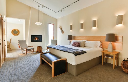 A modern hotel room featuring a bed, seating area, fireplace, and warm lighting in a neutral color palette.