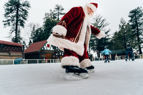 Santa Claus ice skating on a rink, wearing a red suit with white trim, surrounded by trees and other skaters.