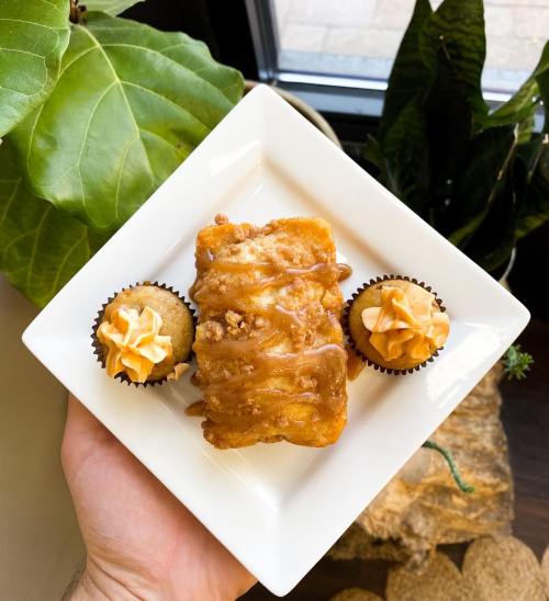 A hand holding a white plate with a glazed pastry and two mini cupcakes topped with orange frosting, surrounded by greenery.