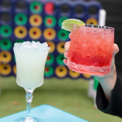 A hand holds a red cocktail with a lime, next to a frosty blue drink in a decorative glass.