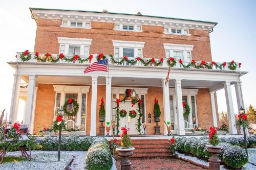 A festive brick house decorated with wreaths and ribbons, featuring an American flag and a snowy front yard.